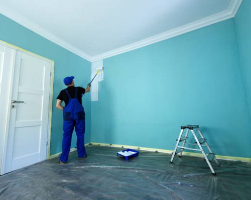 Man in a working overall is painting the wall in white color